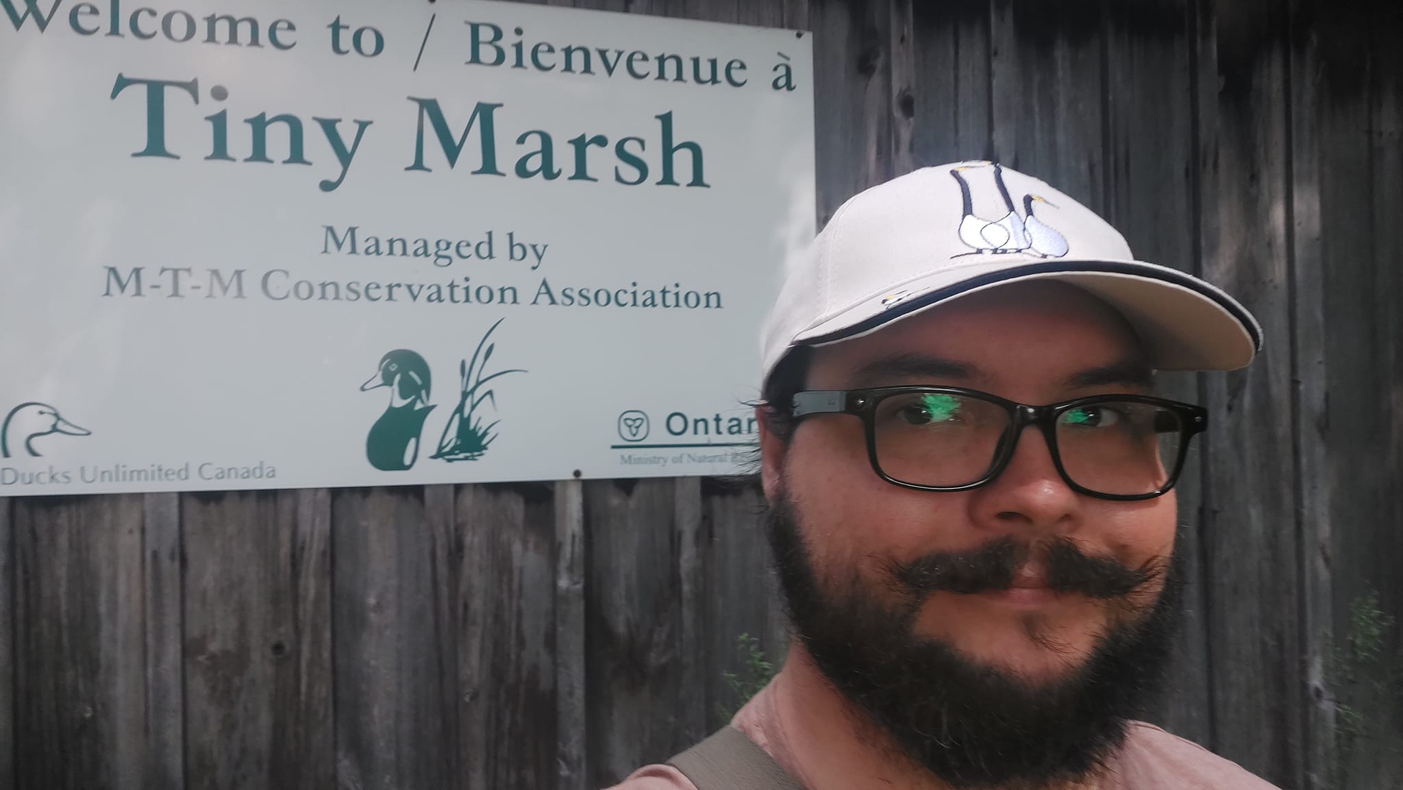 A man short hair and bushy facial hair stands in front of a sign reading "Tiny Marsh" on the side of a weathered wooden building